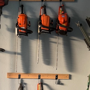 May include: Five orange and black chainsaws hanging on hooks attached to a wooden wall rack. The chainsaws are hanging vertically with the blades facing down.