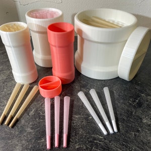 Puede incluir: Un conjunto de seis recipientes de plástico blanco con tapas, algunos de los cuales contienen pequeños palitos blancos. También hay seis palitos blancos y rosados sobre una superficie gris.