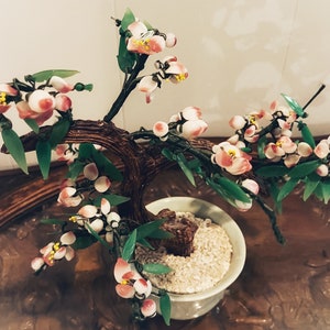 May include: A glass bonsai tree with pink and white flowers and green leaves in a green ceramic pot. The tree is on a wooden surface.