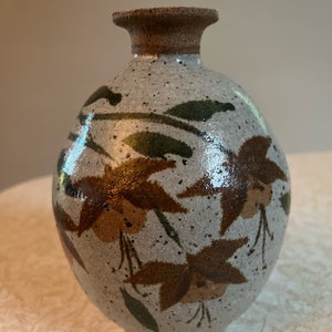 Pode incluir: Um vaso pequeno, redondo, de cerâmica cinza com desenhos florais marrons e verdes. O vaso tem um esmalte salpicado e uma abertura ligeiramente alargada.