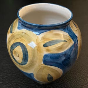 Puede incluir: Un pequeño jarrón redondo de cerámica con un diseño abstracto en azul y amarillo. El jarrón tiene un interior blanco y un borde azul.