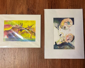 May include: Two framed watercolor paintings of animals. The first painting shows a brown moose with antlers, facing left, with a yellow and green background. The second painting shows a white ram with large curled horns, facing right, with a dark blue background.