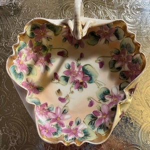 Puede incluir: Un tazón de porcelana blanca con asa, decorado con diseños florales rosas y verdes. El tazón tiene un borde festoneado y está acentuado con ribetes dorados.