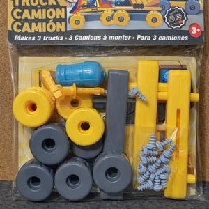 May include: A plastic toy truck construction set in a clear plastic package. The set includes yellow, gray, and blue plastic pieces to build three trucks. The packaging has the text "TRUCK CAMION CAMIÓN" and "Makes 3 trucks".