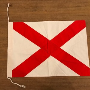 Op de afbeelding: Een witte vlag met een rood diagonaal kruis. De vlag is gemaakt van stof en heeft een touw dat aan de bovenkant is bevestigd.