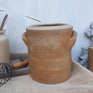 May include: A large, brown stoneware crock with two handles. The crock has a textured surface and is sitting on a beige linen tablecloth.