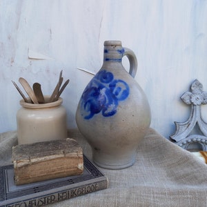 May include: A blue and white stoneware jug with a floral design sits on a hessian surface next to a stack of books. The top book has the title "DECMEISTER ISHIKLOOE" printed on the cover.