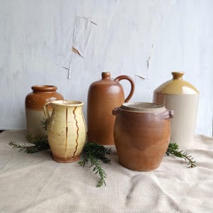 May include: A collection of five ceramic vessels including a tall brown jug with a handle, a cream-colored pitcher with vertical brown drip glaze, two cylindrical jars with two-tone brown and tan finishes, and a smooth tan stoneware bottle.