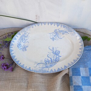 May include: A white oval serving platter with a blue floral design. The platter is on a brown linen tablecloth with a blue and white checkered napkin.