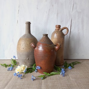 May include: Three antique stoneware vessels in varying shades of brown and tan, including a tall bottle, a wide-mouthed jar with two side handles, and a jug with a single handle, all surrounded by small blue forget-me-not flowers and white blossoms.