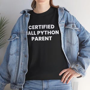 Op de afbeelding: Een zwart t-shirt met de witte tekst CERTIFIED BALL PYTHON PARENT onder een lichtblauw spijkerjack. Het t-shirt heeft een ronde hals en korte mouwen, terwijl het spijkerjack is voorzien van metalen knopen en een klassieke kraag.