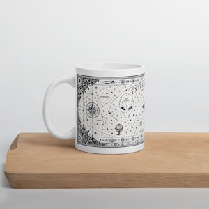 May include: A white ceramic mug featuring a detailed black ink illustration of a celestial map. The design includes constellations, a central alien head, a globe, and the text EXTRA TERRESTRIAL. The intricate border pattern frames the graphic.