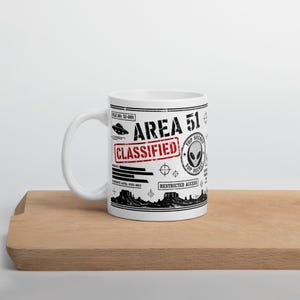 May include: A white ceramic mug featuring black and red graphics with the text AREA 51, CLASSIFIED, and TOP SECRET. The design includes an alien head icon, a flying saucer, desert mountain silhouettes, and various technical document markings.