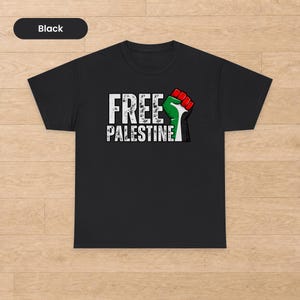 Può includere: Una t-shirt nera a maniche corte con la scritta bianca dall'effetto usurato "FREE PALESTINE" sul petto. Accanto al testo è presente la grafica di un pugno alzato con strisce rosse, verdi, nere e bianche, nei colori della bandiera palestinese.