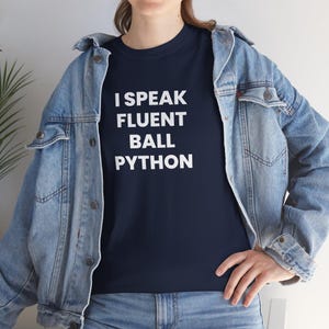 May include: A dark navy blue t-shirt featuring the white text I SPEAK FLUENT BALL PYTHON printed in a bold sans-serif font on the chest, worn underneath a light blue denim jacket with metal buttons and visible stitching details throughout.