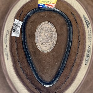 Pode incluir: Um chap&eacute;u de cowboy de couro castanho com uma banda branca e uma faixa de suor de couro preto. O chap&eacute;u tem uma etiqueta que diz "AUSTIN Handmade Hats" e "Custom Handmade". A etiqueta tamb&eacute;m tem uma pequena bandeira americana e o texto "Assembled in USA."