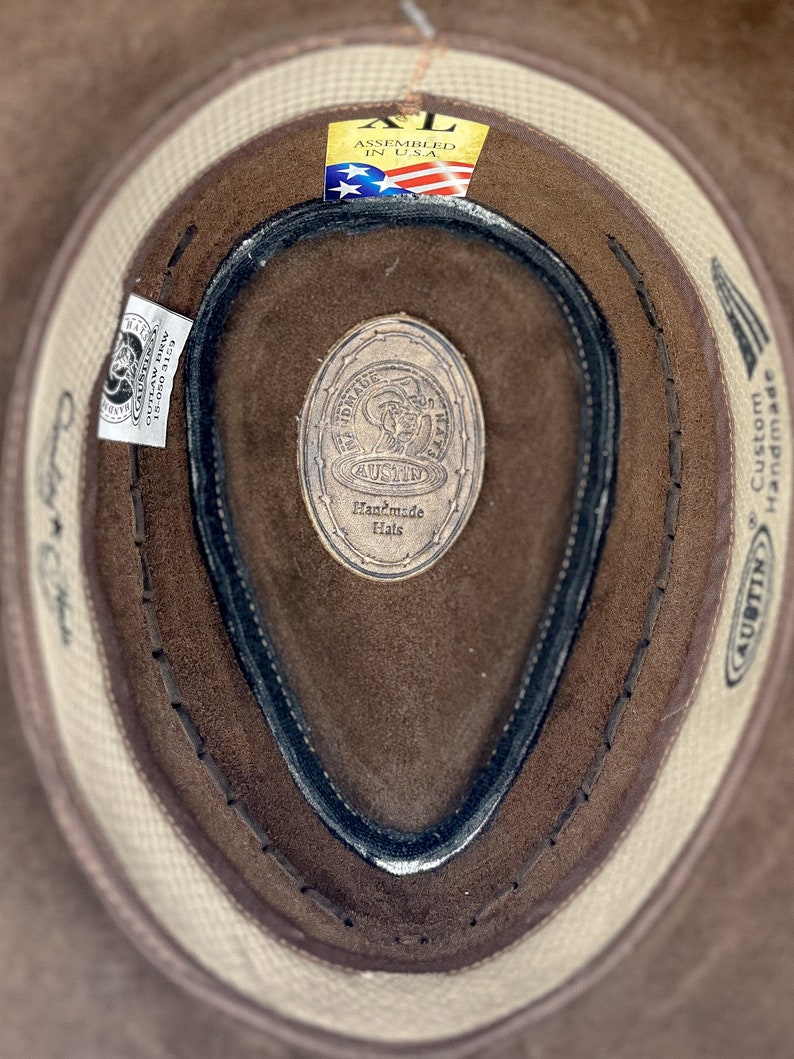 Pode incluir: Um chap&eacute;u de cowboy de couro castanho com uma banda branca e uma faixa de suor de couro preto. O chap&eacute;u tem uma etiqueta que diz "AUSTIN Handmade Hats" e "Custom Handmade". A etiqueta tamb&eacute;m tem uma pequena bandeira americana e o texto "Assembled in USA."
