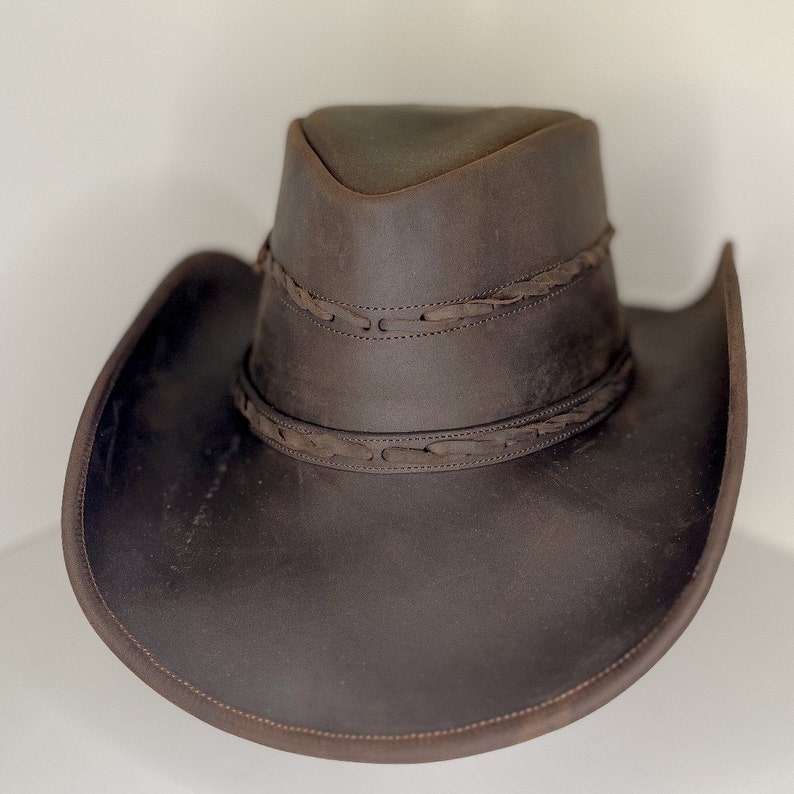 Pode incluir: Um chap&eacute;u de cowboy de couro marrom com uma faixa de couro tran&ccedil;ada ao redor da coroa.