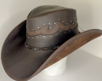 May include: A brown leather cowboy hat with a wide brim. The hat features a decorative band with brass studs and a braided leather band around the crown. The underside of the brim is a lighter brown suede.