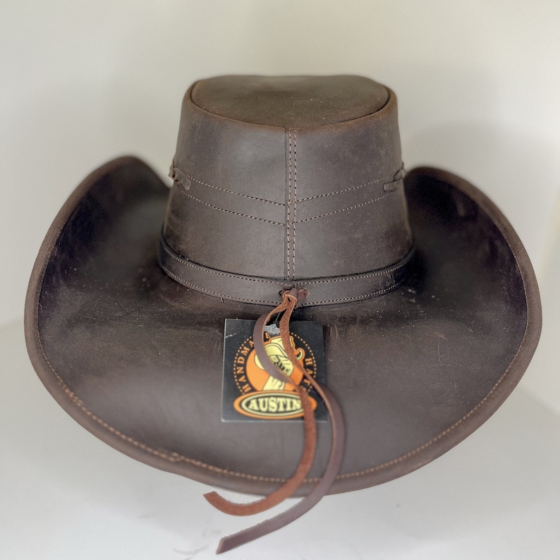 Pode incluir: Um chap&eacute;u de cowboy de couro marrom com uma aba larga e uma faixa de couro. O chap&eacute;u tem uma etiqueta que diz "Handmade Hats Austin".