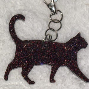 May include: A cat-shaped keychain made of dark material embedded with multi-colored glitter. The silhouette shows a cat in a walking pose with its tail curved upward. A silver-toned metal lobster clasp and split ring are attached to the top.