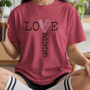 Puede incluir: Una camiseta granate con las palabras "LOVE ERB" impresas en la parte delantera. La palabra "LOVE" tiene un corazón de acuarela que reemplaza la "O". La persona sostiene un vaso de café helado.