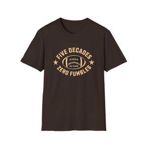 May include: A dark brown t-shirt with a beige graphic. The graphic features a football with the text "FIVE DECADES ZERO FUMBLES" and "UNSTOPPABLE 50 YEARS". Two stars flank the football.
