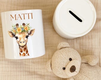 Könnte beinhalten: Weiße Keramik-Spardose mit einem Schlitz oben, neben einem passenden Sockel. Die Spardose zeigt eine Giraffenillustration mit einem Blumenkranz und dem Namen "MATTI" in braunen Buchstaben. Ein beiger Teddybär ist ebenfalls im Bild.
