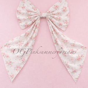 May include: A large fabric bow featuring a white background with a repeating pattern of small pink roses and green leaves. The bow has two long, pointed tails extending downwards and includes the text OGPinksummerdreams printed across the centre.
