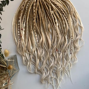 Puede incluir: Dreadlocks rubios y marrón claro con puntas onduladas. Las rastas están atadas con una cinta rosa. Algunas rastas tienen trenzas decorativas con pequeñas cuentas rojas. Las extensiones de cabello están dispuestas en forma de abanico.