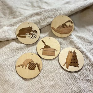 Op de afbeelding: Vijf houten ornamenten met verschillende illustraties van katten. De ornamenten zijn rond en hebben een natuurlijke houten afwerking. De katten zijn afgebeeld in verschillende poses, waaronder spelen met een bol wol, zitten in een doos en klimmen in een kerstboom.