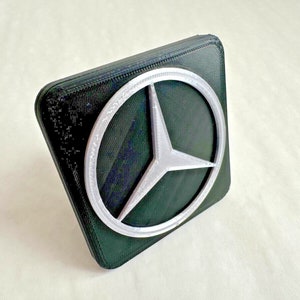 May include: A black square-shaped car emblem with a silver three-pointed star logo.