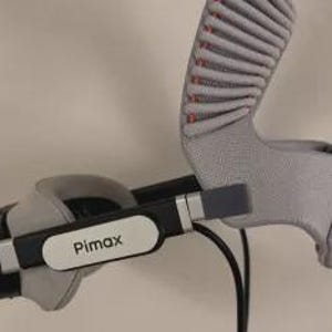 May include: A black and gray virtual reality headset featuring the Pimax logo on the side. The device includes a textured gray head strap with ribbed padding and a black cable extending from the main frame, showcasing a modern design.