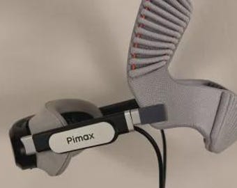 May include: A black and gray virtual reality headset featuring the Pimax logo on the side. The device includes a textured gray head strap with ribbed padding and a black cable extending from the main frame, showcasing a modern design.