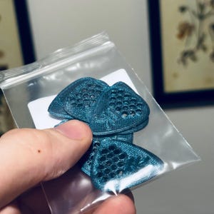 Könnte beinhalten: Nahaufnahme einer Tüte mit tealblauen Gitarrenplektrum. Die Plektrum sind aus einem strukturierten Material gefertigt und haben ein Wabenmuster.