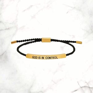 Puede incluir: Una pulsera negra ajustable con detalles dorados. La pulsera presenta la frase "GOD IS IN CONTROL" en letras doradas sobre una barra rectangular. El cordón es negro y los extremos ajustables son dorados.