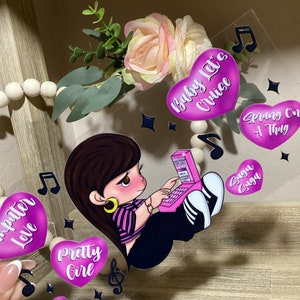 May include: A pink and purple acrylic phone case with a cartoon illustration of a girl with brown hair wearing a striped shirt and black pants. The girl is sitting on a pink phone and typing on a pink laptop. The case has several pink hearts with text on them, including "Baby Let's Cruise", "Spring On A Thug", "Computer Love", "Pretty Girl", "So Fly", and "Suga Suga".