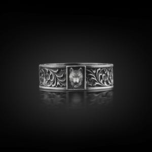 May include: A silver ring with a detailed wolf head centerpiece. The ring features an ornate, swirling floral pattern around the band. The ring is displayed on a reflective black surface.