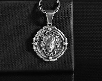 May include: A silver-colored pendant necklace featuring a detailed Medusa head design within a circular frame. The pendant hangs from a textured chain. The necklace is displayed on a black surface, highlighting the intricate details of the jewelry.