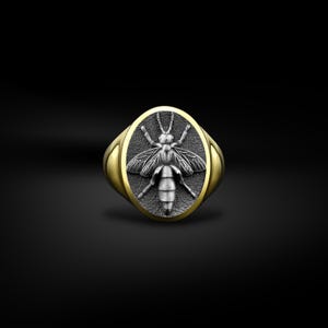 Op de afbeelding: Een goudkleurige ring met een ovale kop met een gedetailleerd, zilverkleurig bijenontwerp op een donkergrijze achtergrond. De ring heeft een gepolijste afwerking. De ring staat tegen een zwarte achtergrond.