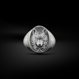 May include: A silver-colored signet ring featuring a detailed wolf head design. The wolf's face is centered within an oval frame, with intricate fur and facial features. The ring has a polished finish and a wide band.