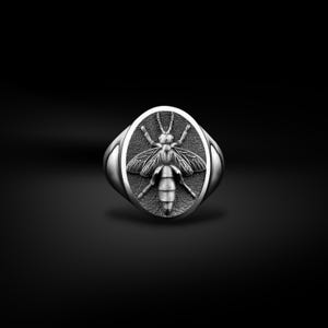 Op de afbeelding: Een zilveren zegelring met een gedetailleerd bijenontwerp. De ovale ring heeft een verhoogd, getextureerd bijenmotief in het midden, met de vleugels en het lichaam van het insect duidelijk gedefinieerd. De ring staat tegen een donkere achtergrond.