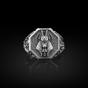 Puede incluir: Anillo plateado con diseño de Anubis. El anillo tiene una cara octogonal con una representación detallada de Anubis, el dios egipcio del más allá, y una calavera. El anillo tiene un borde adornado con motivos florales.