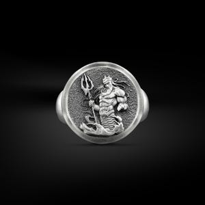 Pode incluir: Anel de prata com um relevo detalhado de uma figura musculosa e barbuda segurando um tridente. A figura é retratada emergindo das ondas, usando uma coroa e está emoldurada em um círculo. O anel tem um acabamento polido.