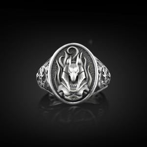 Puede incluir: Anillo plateado con un diseño ovalado de Anubis, con llamas y una luna creciente. El anillo tiene detalles laterales intrincados y un acabado pulido, sobre un fondo oscuro.