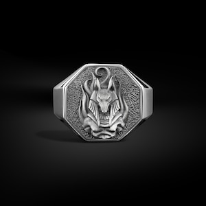 Puede incluir: Anillo plateado con una cara octogonal que presenta un relieve detallado de Anubis, el dios egipcio del más allá, con llamas alrededor de su cabeza. El anillo tiene un fondo texturizado y un acabado pulido.