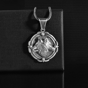 May include: Silver-toned pendant necklace featuring a detailed wolf head design within a circular frame. The pendant hangs from a matching chain, set against a dark, textured background. A striking piece of jewelry.
