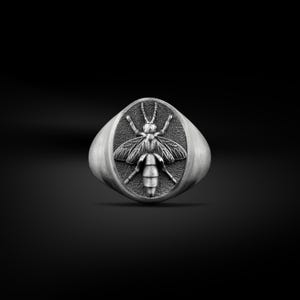 Op de afbeelding: Een zilverkleurige ring met een gedetailleerd bijenontwerp. De bij is gecentreerd op een ovaalvormig vlak, met ingewikkelde vleugel- en lichaamsdetails. De ring heeft een geborstelde metalen afwerking, tegen een donkere achtergrond.