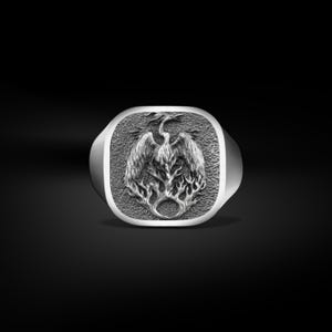 May include: A silver ring with a square face featuring a detailed relief of a phoenix rising from flames. The bird's wings are spread, and the design is set against a textured background. The ring has a polished finish.