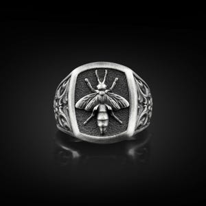 Op de afbeelding: Zilverkleurige ring met een gedetailleerd bijenontwerp. De bij is gecentreerd op een rechthoekige plaat, omlijst door sierlijke krullen. De ring heeft een gepolijste afwerking, tegen een donkere achtergrond.